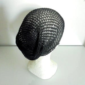 Pode incluir: Um gorro preto de tecido aberto exibido em uma cabeça de manequim branca. A textura do gorro é visível, com um padrão solto semelhante a uma rede. O gorro é de cor escura e sólida, e o estilo é casual.
