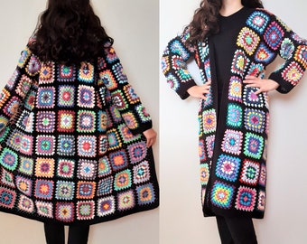 Könnte beinhalten: Ein langer, schwarzer Häkelcardigan mit einem bunten Patchwork-Muster. Der Cardigan besteht aus Granny Squares in verschiedenen Farbtönen von Rosa, Blau, Grün, Gelb und Lila.