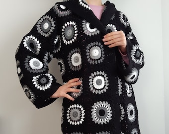 May include: A black crochet cardigan with a hood, featuring a circular pattern in shades of white and gray.
