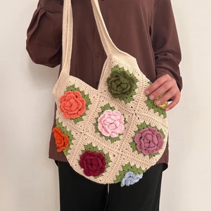May include: A cream-colored crochet tote bag adorned with colorful, three-dimensional floral appliqués. The bag features a square pattern design with roses in shades of orange, pink, red, green, and blue. The bag has a long strap for shoulder or hand carry.