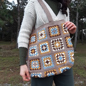 Op de afbeelding: Handgemaakte gehaakte tote bag met een patchwork ontwerp. De tas is voornamelijk bruin met vierkante patches in blauw, geel en crème. Het heeft een bijpassende bruine band. Ideaal voor boodschappen of dagelijks gebruik.