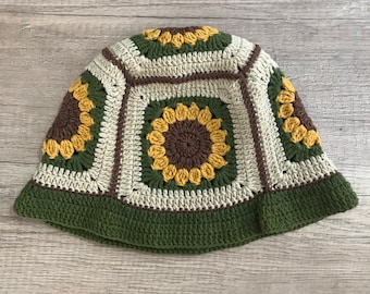 Puede incluir: Un gorro de cubo tejido a crochet con un patrón de girasol en tonos marrones, amarillos y verdes.