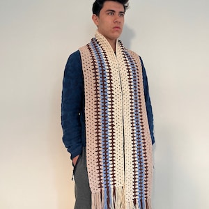May include: A crocheted scarf with brown, white, and blue stripes and fringe. The scarf is worn by a person standing in front of a white wall.