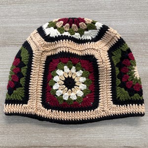 May include: A handmade crochet beanie hat with a square pattern. The hat features a central square with a floral design in black, white, and burgundy. The surrounding squares are in black, green, and beige. The brim is black.
