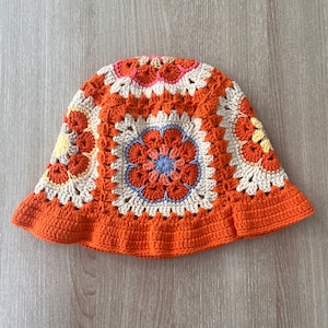 Op de afbeelding: Een gehaakte bucket hat met een kleurrijk bloemenpatroon in tinten oranje, roze, geel en blauw. De hoed heeft een brede rand en een gehaakt ontwerp.