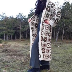 Könnte beinhalten: Ein langer weißer Häkelcardigan mit Kapuze, der ein geometrisches Muster aus braunen und grünen Kreisen aufweist. Der Cardigan wird über einem dunkelgrauen Oberteil und einer Hose getragen.