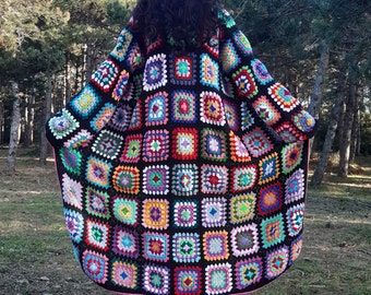 Könnte beinhalten: Ein langer, schwarzer Häkelcardigan mit einem bunten Granny-Square-Muster. Der Cardigan wird von einer Person getragen, die in einem Wald steht.