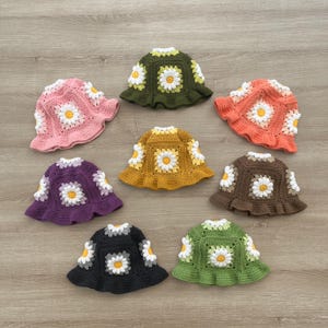 Op de afbeelding: Een verzameling van acht handgemaakte gehaakte emmerhoeden in verschillende kleuren, waaronder roze, groen, oranje, paars, geel, bruin, zwart en lichtgroen. Elke hoed heeft een madeliefjesbloemmotief.