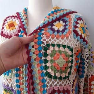 Op de afbeelding: Een kleurrijk gehaakt vest met een granny square patroon in tinten rood, oranje, geel, groen, blauw en paars. Het vest is gemaakt met wit garen en heeft een losse, open structuur.