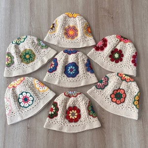 Puede incluir: Siete sombreros de cubo de ganchillo en varios colores con patrones florales. Los sombreros están hechos con hilo beige y presentan flores coloridas en tonos de rosa, azul, verde, rojo y amarillo.