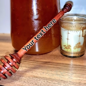 Può includere: Un mestolo di legno per il miele con una finitura rosso-marrone e un design a spirale. Il mestolo è appoggiato su una superficie di legno accanto a un barattolo di vetro di miele e una tazza di vetro con un design dorato.