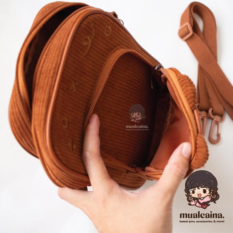 Op de afbeelding: Een bruine corduroy crossbodytas met een hoofdvak met ritssluiting en een verstelbare schouderriem. De tas heeft een rechthoekige vorm met afgeronde hoeken en een contrasterende bruine rand. De binnenkant is zichtbaar en de tas is open.