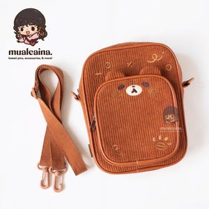 Op de afbeelding: Bruine corduroy crossbodytas met een berengezichtontwerp op de voorzak. De tas heeft een lange, verstelbare riem en een pootafdrukdetail. De merknaam "mualcaina" is zichtbaar.