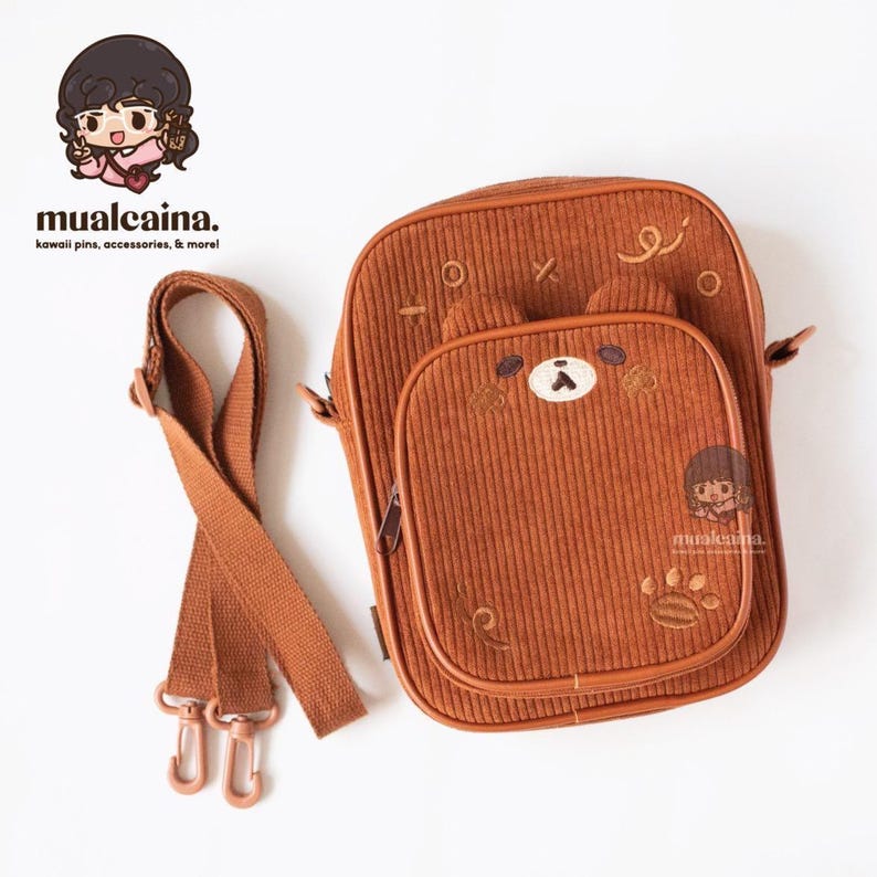 Op de afbeelding: Bruine corduroy crossbodytas met een berengezichtontwerp op de voorzak. De tas heeft een lange, verstelbare riem en een pootafdrukdetail. De merknaam "mualcaina" is zichtbaar.