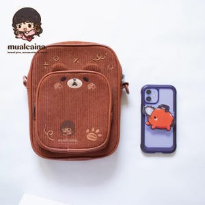 Op de afbeelding: Bruine corduroy crossbodytas met een berengezichtontwerp op de voorzak. Naast de tas ligt een paarse telefoonhoes met een oranje kettingzaagkarakter. De tas heeft de tekst "mualcaina" en andere kawaii-accessoires.