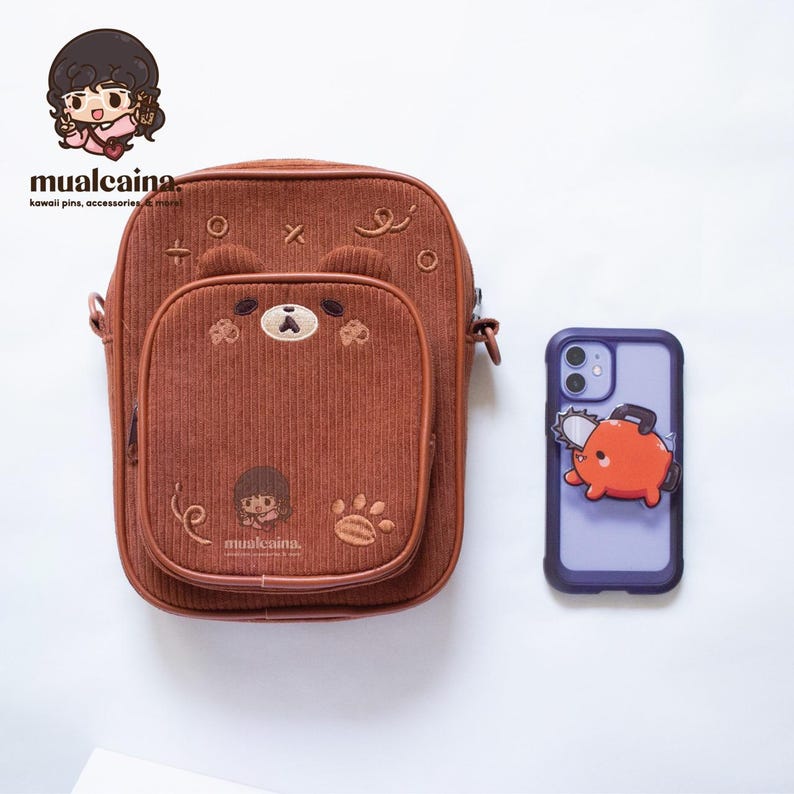 Op de afbeelding: Bruine corduroy crossbodytas met een berengezichtontwerp op de voorzak. Naast de tas ligt een paarse telefoonhoes met een oranje kettingzaagkarakter. De tas heeft de tekst "mualcaina" en andere kawaii-accessoires.