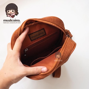 Op de afbeelding: Open zicht op een bruine corduroy crossbody tas. De tas heeft een ritssluiting en een binnenvak. De merknaam "mualcaina" is zichtbaar op een label in de tas. Witte achtergrond.