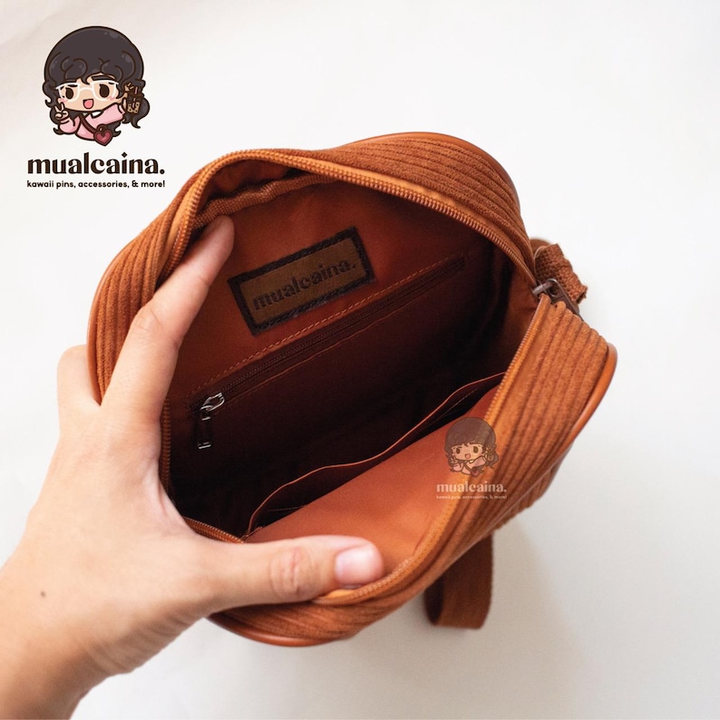 Op de afbeelding: Open zicht op een bruine corduroy crossbody tas. De tas heeft een ritssluiting en een binnenvak. De merknaam "mualcaina" is zichtbaar op een label in de tas. Witte achtergrond.
