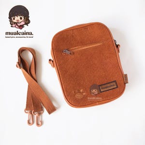 Op de afbeelding: Een bruine corduroy crossbody tas met ritssluiting en verstelbare riem. De tas heeft een klein logo en een cartoon karakter ontwerp. Ontworpen voor het dragen van persoonlijke spullen.