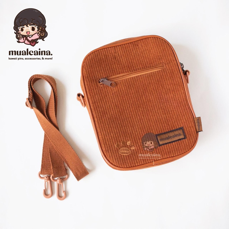 Op de afbeelding: Een bruine corduroy crossbody tas met ritssluiting en verstelbare riem. De tas heeft een klein logo en een cartoon karakter ontwerp. Ontworpen voor het dragen van persoonlijke spullen.