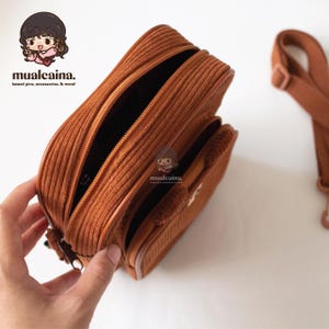 Op de afbeelding: Een bruine corduroy crossbody tas met een hoofdvak met ritssluiting en een kleinere voorzak. De tas heeft een bijpassende verstelbare riem. De tekst "mualcaina. kawaii pins, accessories, & more!" is zichtbaar.