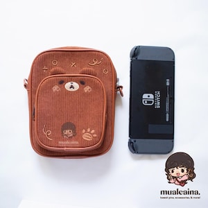 Op de afbeelding: Een bruine corduroy crossbodytas met een berengezichtontwerp en een Nintendo Switch-console. De tas heeft een geborduurd berengezicht, een klein vak en de tekst "mualcaina."