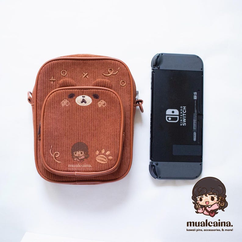 Op de afbeelding: Een bruine corduroy crossbodytas met een berengezichtontwerp en een Nintendo Switch-console. De tas heeft een geborduurd berengezicht, een klein vak en de tekst "mualcaina."