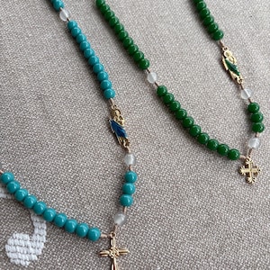 May include: Two rosaries featuring turquoise and green beads. The rosaries have gold-coloured crosses and decorative elements. Clear and white beads are interspersed. The rosaries are displayed on a textured, neutral-coloured surface.