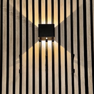 May include: A modern black and gold wall sconce with two light sources, mounted on a vertical wood slat wall. The sconce casts a warm glow, and the wall features alternating light wood and black stripes. Two black hooks are also visible.