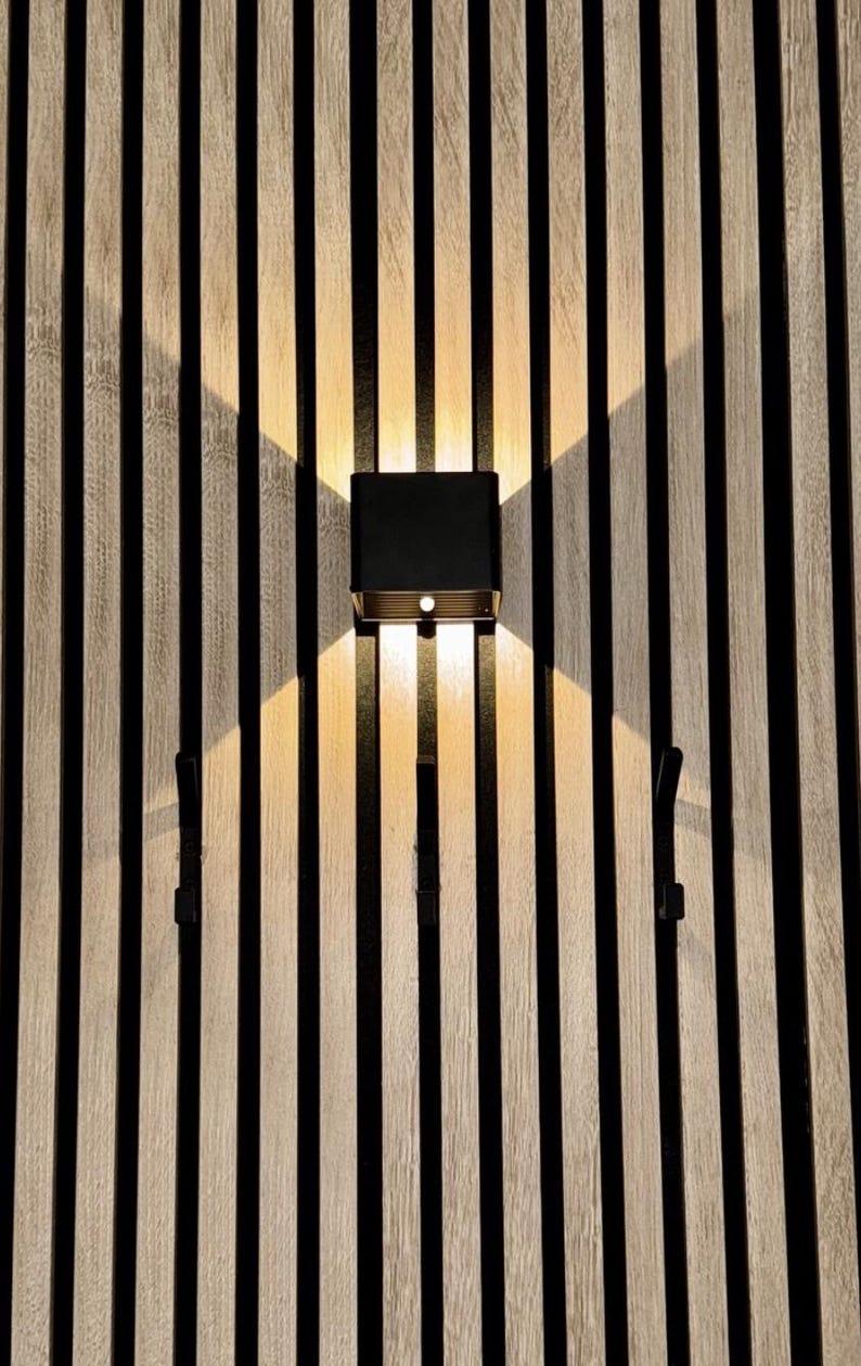 May include: A modern black and gold wall sconce with two light sources, mounted on a vertical wood slat wall. The sconce casts a warm glow, and the wall features alternating light wood and black stripes. Two black hooks are also visible.