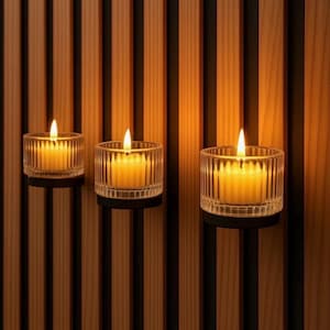 Op de afbeelding: Drie brandende kaarsen in heldere, geribbelde glazen houders. De kaarsen staan tegen een achtergrond van verticale houten latten in bruin- en zwarttinten. De warme gloed van de vlammen creëert een gezellige sfeer.