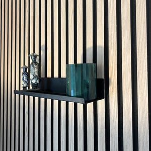 Op de afbeelding: Een zwart metalen plank met twee kleine keramische beeldjes en een groene glazen vaas. De plank is gemonteerd aan een muur met verticale houten panelen.