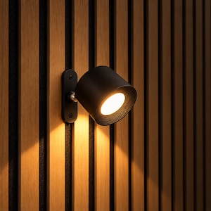 Könnte beinhalten: Eine mattschwarze zylindrische Wandleuchte mit runder Montageplatte und Drehgelenk. Die Leuchte verfügt über einen gerippten Innenrand und strahlt ein warmes, helles Licht aus, das die umliegenden vertikalen Holzlatten beleuchtet.