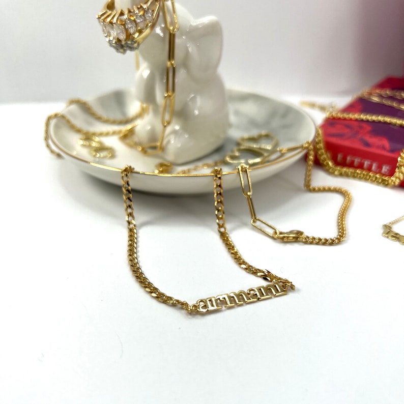 A custom made to order gold name bracelet displaying the name Armani lays out over a white ceramic elephant thats displayed on a white background. Also laid out in white elephants is a variety of gold jewelry made by Jaelyn Jewelry. Name bracelet.