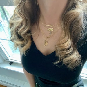 May include: A woman wearing a black v-neck shirt with a layered gold necklace. The necklace features the words "worthy", "1986", and the letter "J".