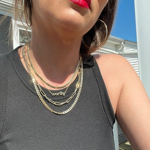 May include: A woman wearing a black tank top with a layered gold chain necklace. The necklace features a small pendant with the word "worthy" on it. She is also wearing large gold hoop earrings and sunglasses.