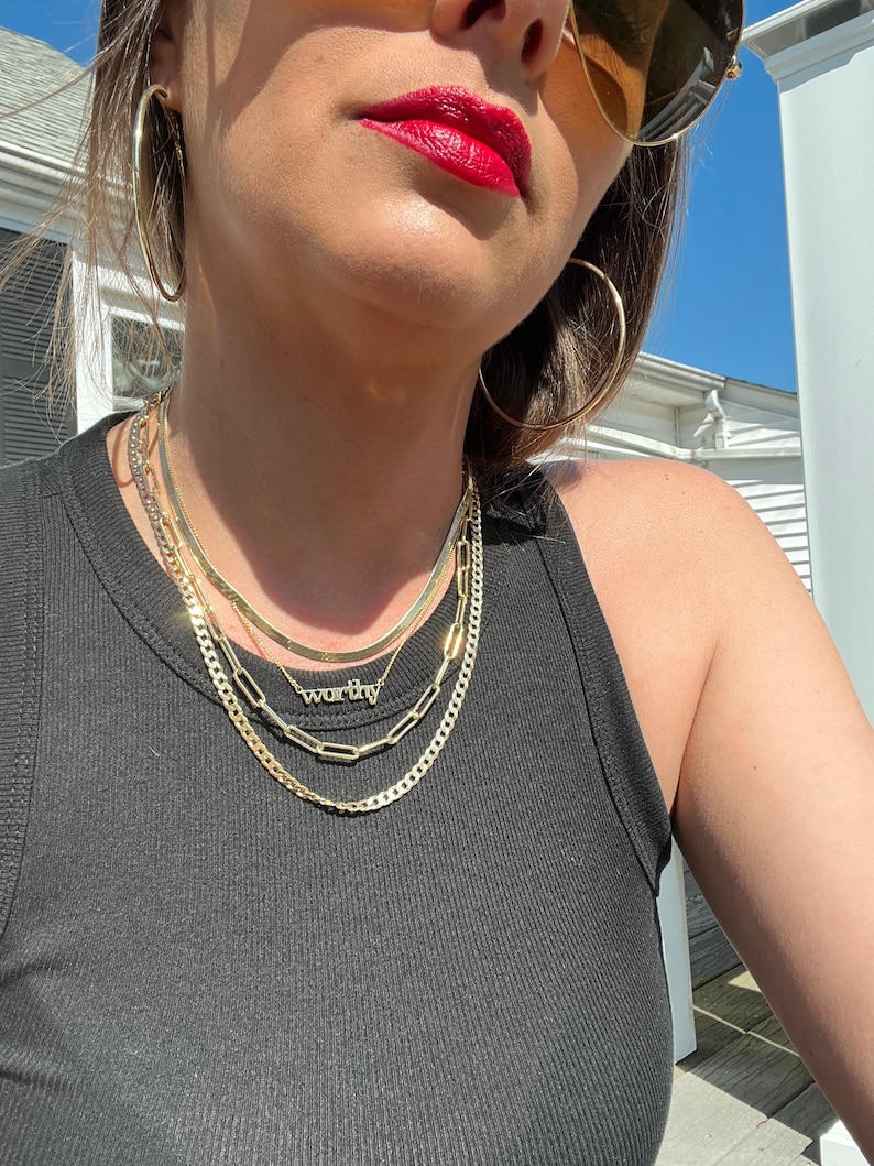 May include: A woman wearing a black tank top with a layered gold chain necklace. The necklace features a small pendant with the word "worthy" on it. She is also wearing large gold hoop earrings and sunglasses.