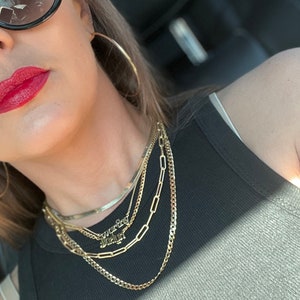 May include: A woman wearing a black tank top and gold necklaces. The necklaces include a thin chain, a thicker chain, a chain with rectangular links, and a nameplate necklace that says "worthy" and "enough".