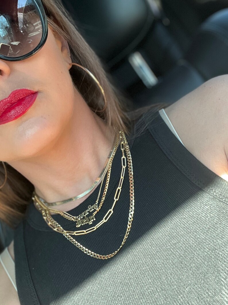 May include: A woman wearing a black tank top and gold necklaces. The necklaces include a thin chain, a thicker chain, a chain with rectangular links, and a nameplate necklace that says "worthy" and "enough".