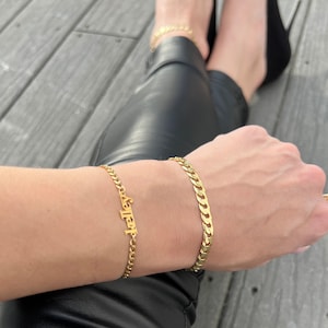 A woman wearing black leather legging pants and pointed black stilettos sits on a grey flooring with her legs in a kriss Kross while laying her arm over her pants and displaying her 2 gold bracelets made by Jaelyn Jewelry Co. Gold name bracelet. Curb