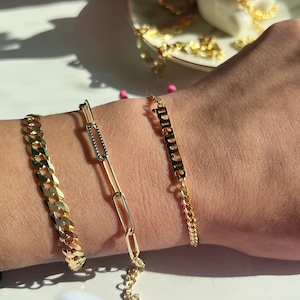 A young woman lays her wrist out and displays her 3 gold bracelets made by Jaelyn Jewelry Co. She is seen wearing a personalized chunky gold name bracelet that's displays the name Armani. Gold curb chain bracelet and a paperclip bracelet.