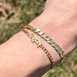 A woman stands outside on a bright sunny day and models her 2 gold bracelets over a green grass backdrop with the bright sun reflecting off of the precious gold. She is wearing a curb chain bracelet and a custom made gold lowercase name bracelet.