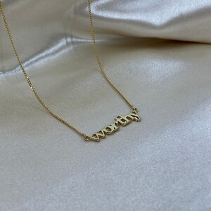 May include: A gold chain necklace with the word "worthy" spelled out in gold letters.