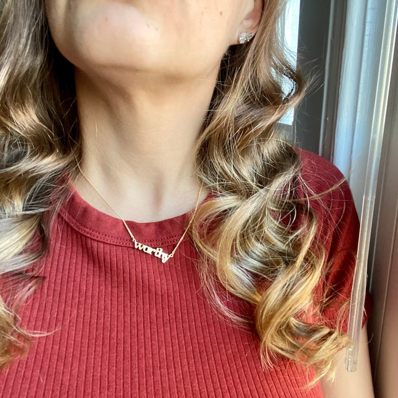 May include: A woman wearing a red shirt with a gold necklace that says "worthy".