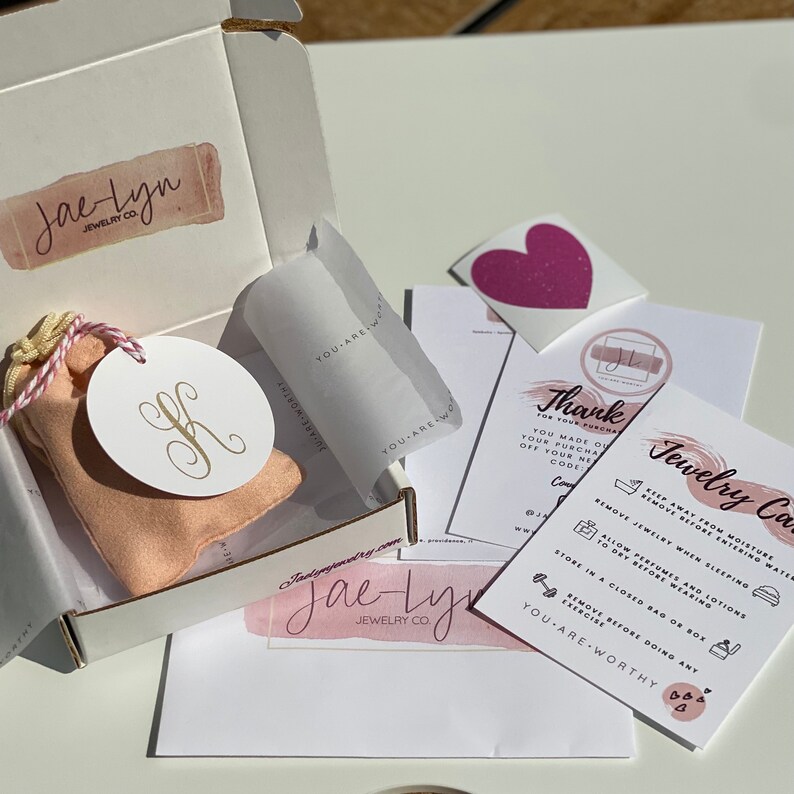 May include: A pink and white gift box with a pink heart sticker and a card that says "Thank you for your purchase. You made our day!" and "Jewelry Care: Keep away from moisture, water, and oils. Remove jewelry when sleeping. Allow perfumes and lotions to dry before wearing. Store in a closed bag or box. Remove before doing any exercise. You are worthy."