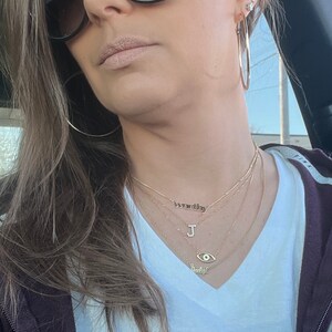 May include: A woman wearing a white t-shirt with a gold necklace that says "worthy" and a gold necklace with a letter "J" charm. She is also wearing a gold necklace with an evil eye charm and a gold necklace that says "babygirl".