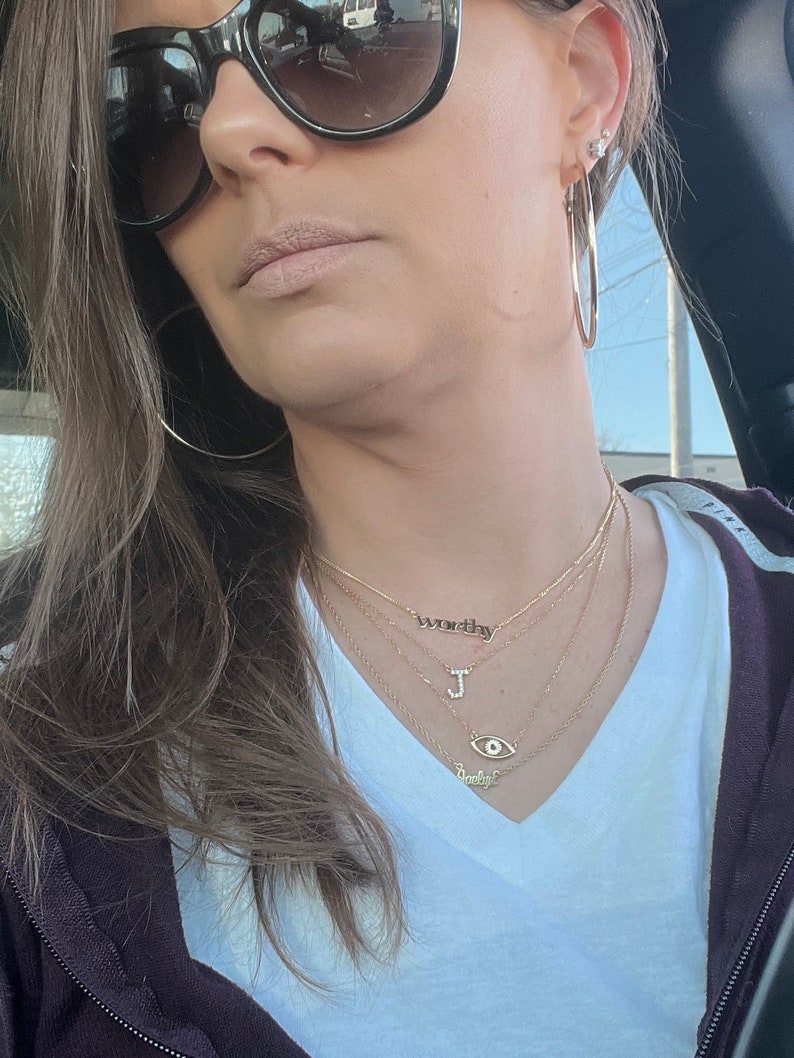 May include: A woman wearing a white t-shirt with a gold necklace that says "worthy" and a gold necklace with a letter "J" charm. She is also wearing a gold necklace with an evil eye charm and a gold necklace that says "babygirl".