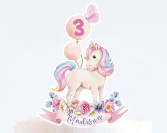 May include: A pink and white unicorn cake topper with a pink balloon that says "3" and a pink butterfly. The unicorn is surrounded by pink and purple flowers and a banner that says "Madisun".