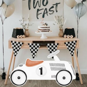 May include: A white toy car with a brown seat and black and white checkered details. The car has a number "1" on the side. The car is in front of a table with a cake and balloons. The balloons are black and white checkered, light blue, and beige. The table has a sign that says "Two Fast".