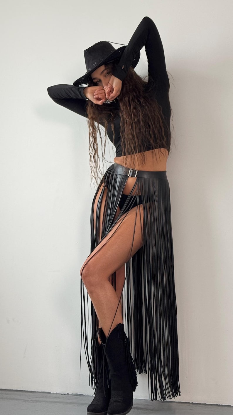 Puede incluir: Una falda de flecos de cuero negro con cintur&oacute;n y hebilla. La falda es larga y fluida, con los flecos que llegan al suelo. La modelo lleva una camisa negra de manga larga y un sombrero vaquero negro.